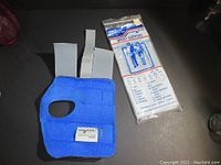 Blue adjustable wrist support with three grey straps next to its original clear packaging with blue and red text.