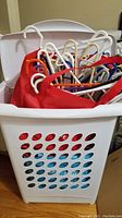 White Sterilite plastic laundry hamper with visible ventilation holes loaded with assorted colored plastic hangers inside, red cloth is partially inside