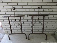 Two brown metal folding table legs shown side by side against a white brick wall. Both are upright with visible mounting plates on top and folding hinges.