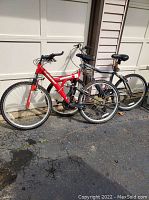 Two bicycles side by side outdoors on pavement against a garage door background – one red and one gray, both mountain bikes with dual suspension.