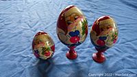 Three vintage handmade wooden Santa Claus eggs placed on blue cloth background: one small without stand, one medium and one large on red stands