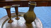 Photo showing all items: a large copper tray, two brass candle holders, and one brass vase on a blue cloth background.
