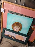 Various framed wall art, including a portrait painting of a woman, an embroidered floral piece, and others stacked together.