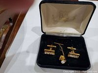 Photo shows the pair of gold-tone cufflinks with multiple embedded clear stones and the tie pin with a chain inside the black carrying case with plush interior.