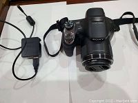 Sony Cyber-Shot DSC-H400 camera with charger and attached shoulder strap shown from above on a white background.