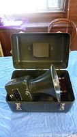 Photo of vintage military super hailer unit inside its carrying case, top lid open, showing olive green metal construction and horn