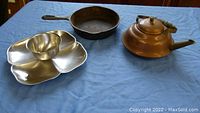 Photo showing the copper/brass teapot, stainless steel chip and dip tray, and cast iron skillet laid out on a blue cloth.