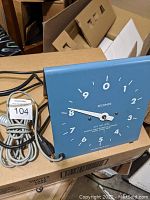 Front side view of blue square electronic stop clock showing second dial and hands with power cord coiled beside the unit.