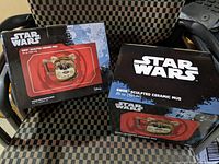 Two boxed Star Wars EWOK sculpted ceramic mugs shown side by side on a checkered chair, boxes intact and labeled.
