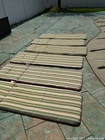 Six green and white striped pool lounger cushions laid out side by side outdoors on a stone patio showing overall condition and pattern.
