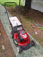 Red Craftsman Gold lawn mower with a beige grass collection bag, situated outdoors on a paved path beside some soil and vegetation.