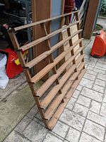 Photo showing custom wooden display rack with angled horizontal slats, standing on floor outside a garage.