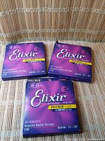 Three packs of Elixir Polyweb Acoustic Guitar Strings in medium gauge .013-.056, 80/20 Bronze, displayed on a surface.