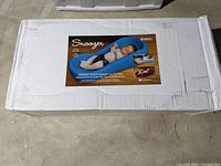 Box showing the product image and description indicating the Keekaroo Snoozer infant stationary sleeper with a baby lying on the gray cushioned lounge.