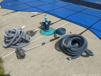 Various pool cleaning equipment including flexible hoses, brushes, and vacuum heads arranged on poolside concrete near covered pool.