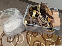 Belt collection in plastic bin with various belts visible including wide leather and metallic belts, some wrapped in paper.