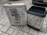 Photo of the Saunatec electric sauna heater next to the original packaging box labeled 'The Original Finnish Sauna', showing the heater's metallic housing and top heating elements.