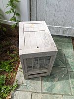 Rectangular beige plastic hose reel housing with wood panel texture, sitting on tiled ground next to a garden bed.