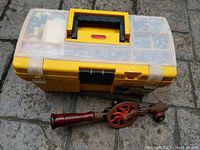 Closed yellow plastic toolbox with black handle and vintage wooden-handled hand drill lying next to it on ground