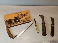 Full view of all four items: Hostess Frozen Food Saw in original box, small yellow pocket knife, two curved cutting tools with wooden handles.
