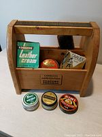 Wooden shoe caddy with ribbed top and brush, holding vintage leather cream, boot polish tins and carton boxes in front and inside compartments.