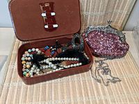 Open brown leather jewelry box with various necklaces including pearl and beaded styles, and a pair of large black earrings inside. Pink sequin clutch bag with silver frame and chain strap next to it.