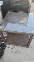 View of square patio table showing clear glass top with metallic corner fasteners and rattan woven base with lower shelf.