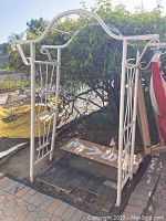 Front and side view of white metal outdoor garden archway with scrollwork and lattice sides amid greenery.