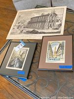 Three art prints laid on a table: one large architectural lithograph print, one framed etching of a gondolier, and one framed lithograph with an architectural theme.