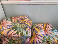 Three folded patio chair cushions with tropical leaf pattern stacked.
