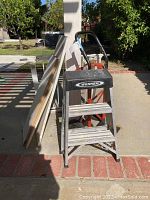 Photo showing Werner two-step aluminum ladder, orange metal dolly, and wooden shelves leaning against a post outdoors.