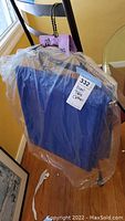 Two blue rectangular event table cloths folded and wrapped in plastic, hanging over a chair in a sealed wrapping with a visible label.