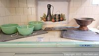 Three Fire King jadeite green nesting bowls in small, medium, large sizes arranged together on a tiled countertop near a colander and placemats.