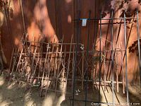 Multiple iron fence sections leaning against a wall casting shadows, showing pointed spear finials and scrollwork, with visible rust and surface corrosion.