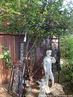 Full view of the iron garden arch with two decorative iron panels leaning against it, placed outside with garden plants around. A statue stands beside but is not included in the lot.