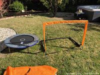 Small round trampoline and orange framed soccer goal net on grass outdoors
