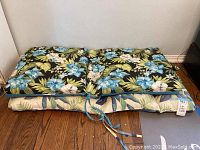 Two patio lounge chair cushions stacked, showing the top cushion with a dark floral pattern and blue ties.