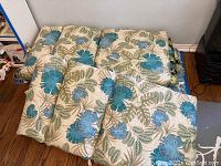 Six patio lounge chair cushions stacked next to a wall, showing the blue floral and green leafy pattern.