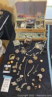 Full display of vintage costume jewelry pieces laid out on a black cloth with open multi-compartment jewelry storage box in background, containing additional jewelry items and rings.