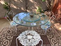 Patio set including glass oval table top on iron base, two iron chairs with cushions, and iron decorative stool placed on outdoor rug.