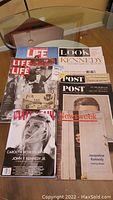 Photo showing a collection of vintage magazines on a glass table, featuring Life, Look, Post, Newsweek, and Vanity Fair magazines, many with covers related to John F. Kennedy and his family.