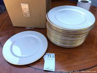 Stack of 20 white plates with gold trim on a wooden table, alongside a cardboard box and MaxSold lot tag