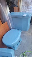Photo showing powder blue toilet tank with lid and matching toilet bowl with seat down placed on ground indoors near other items.
