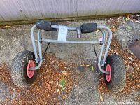 Side view of the boat trolley showing the metal frame, foam rollers, and large pneumatic tires with red hubs on a paved surface with leaves and dirt, evidencing outdoor use and wear.