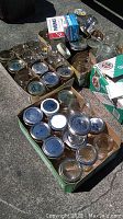 Multiple clear glass canning jars arranged in two boxes, most have metal lids, some jars without lids, shown outdoors on pavement.
