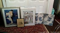 Front view of five artwork pieces and fabric panels, showing three framed signed artwork and two unframed fabric panels with blue floral patterns, all displayed against a white door on a red patterned carpet.