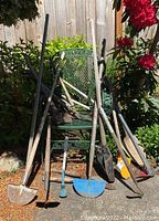 Photo showing various long-handled gardening and yard tools arranged on and leaning against a green metal garden chair outdoors. Tools include shovels, hoes, an axe, and other digging tools with wooden handles and rusted metal parts.