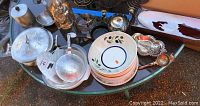 Overview of multiple metal and aluminum kitchenware items on table including round lacquered trays, bowls, plates, flatware and serving pieces.