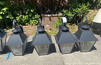 Four black metal and glass outdoor wall lanterns with mounting hooks placed outdoors on pavement in sunlight.