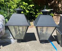 Pair of black metal outdoor lamp post lights with textured glass panels shown outdoors on pavement with greenery background.